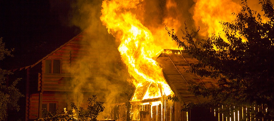 Burning wooden house at night. Bright orange flames and dense smoke from under the tiled roof on dark sky, trees silhouettes and residential neighbor cottage background. Disaster and danger concept.